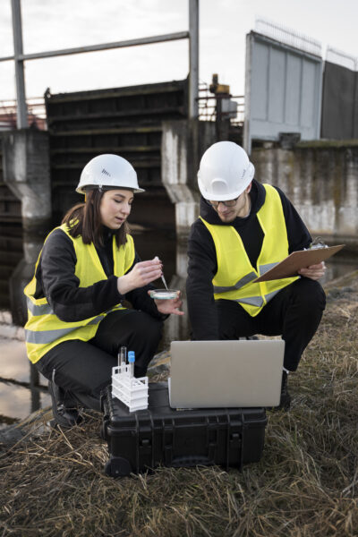 A Empresa 2 Dois engenheiros usando capacetes brancos e coletes de segurança amarelos, um homem e uma mulher, estão conduzindo testes de qualidade da água em um local industrial ao ar livre. A engenheira está testando uma amostra de água enquanto o engenheiro anota observações em uma prancheta, provavelmente para um estudo ambiental, com um laptop por perto.