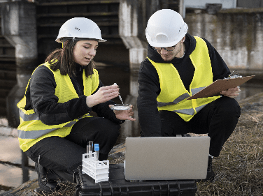 Dois engenheiros usando coletes de segurança amarelos e capacetes brancos estão trabalhando ao ar livre. Um está segurando uma amostra de teste e uma pipeta, enquanto o outro está apontando para uma tela de laptop. Uma prancheta e tubos de ensaio também são visíveis, pois eles garantem o licenciamento ambiental perto de uma estrutura industrial.